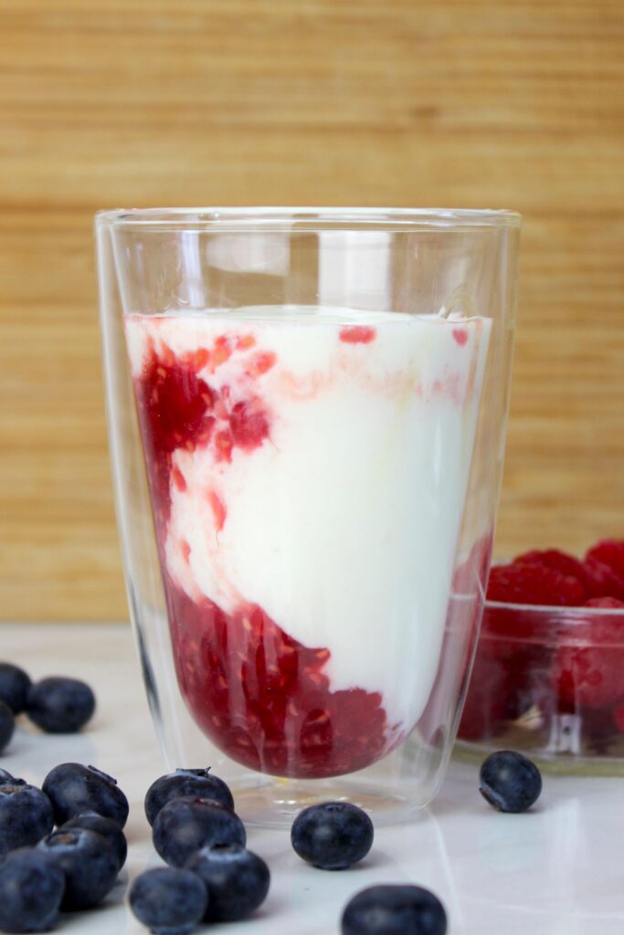 Himbeer-Kefir-Drink im Glas vor Holzuntergrund, mit sichtbarer roter Fruchtschicht und umgeben von frischen Blaubeeren und Himbeeren