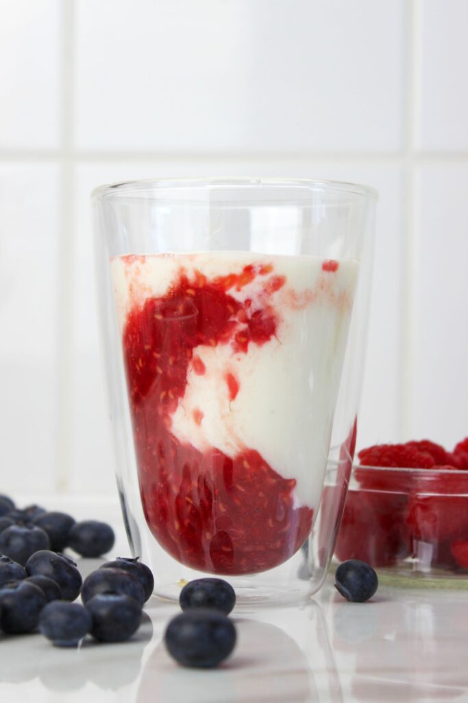 Himbeer-Kefir-Drink im doppelwandigen Glas mit sichtbarer Fruchtschicht, daneben frische Heidelbeeren und Himbeeren auf weißem Untergrund.