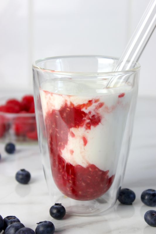 Himbeer-Kefir-Drink im doppelwandigen Glas mit sichtbarer Fruchtschicht, daneben frische Heidelbeeren und Himbeeren auf weißem Untergrund.
