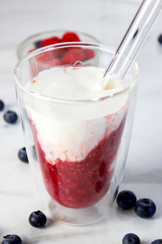 Himbeer-Kefir-Drink im doppelwandigen Glas mit sichtbarer Fruchtschicht, daneben frische Heidelbeeren und Himbeeren auf weißem Untergrund.
