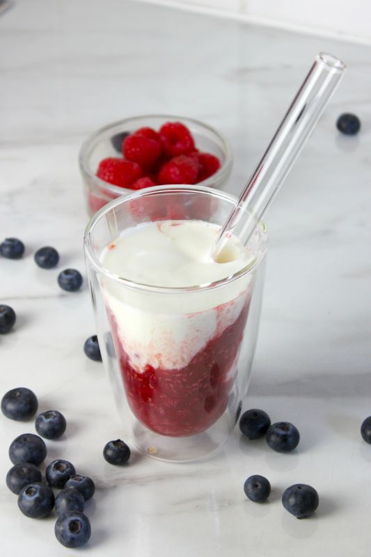 Himbeer-Kefir-Drink im doppelwandigen Glas mit sichtbarer Fruchtschicht, daneben frische Heidelbeeren und Himbeeren auf weißem Untergrund.